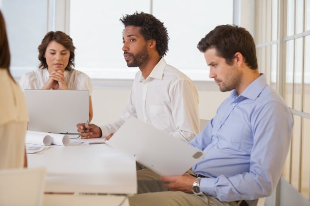 An American business leader is sitting with their team during a meeting. They are all reviewing documents. The leader looks at the team, directly engaging with them on their ideas and challenges.