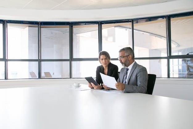 Two US businesspeople sitting at a conference table, reviewing a project plan. They appear to be discussing strategies, and one is gesturing with a pen, emphasizing a point. The atmosphere is collaborative and focused.